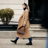 Mujer con abrigo camel, bolso marrón y mocasines negros, un look de quiet luxury paseando por la ciudad.