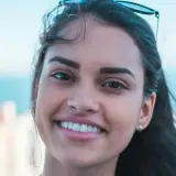 Sonrisa radiante de una joven con rostro ovalado, cabello oscuro y gafas de sol en la cabeza. Fondo de ciudad y mar.