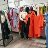 Dos mujeres discuten el outfit significado de un vestido naranja vibrante en una boutique.