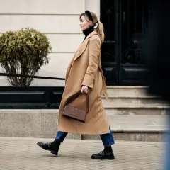 Mujer con abrigo camel, bolso marrón y mocasines negros, un look de quiet luxury paseando por la ciudad.