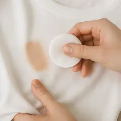 Mano limpiando una mancha de maquillaje de una camiseta blanca con un disco de algodón. Guía rápida para como quitar maquillaje de la ropa.