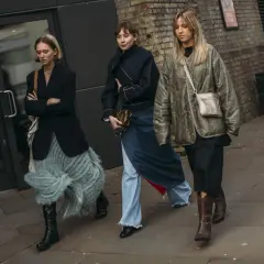 Tres mujeres caminan por la calle, cada una con un estilo único. La de la izquierda lleva unas **botas** negras altas y una falda de flecos. La del medio, un conjunto vaquero y unas botas negras. La de la derecha, unas botas marrones y una chaqueta aco...