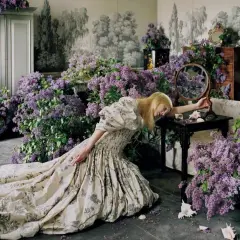 Una mujer rubia con un vestido estampado cae sobre un montón de lilas, evocando la estética onírica de Tim Walker.