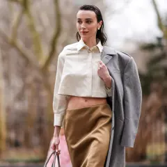 Mujer con blusa corta y falda satinada color caramelo, abrigo gris colgado del hombro.