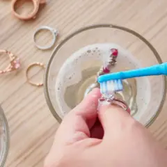 Manos limpian joyas con un cepillo azul en agua con espuma. Otros anillos esperan su turno.