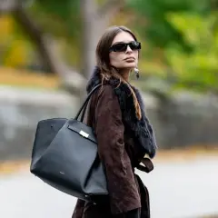 Mujer con abrigo marrón y bolso negro de Demellier, gafas de sol y pendientes llamativos.
