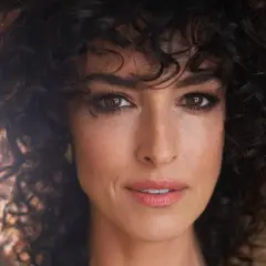 Primer plano de una mujer con un flequillo cortina de pelo rizado oscuro, sus ojos marrones miran directamente a la cámara.