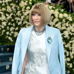 Anna Wintour, figura clave de que es la Gala Met, posa con un traje azul claro y un vestido blanco bordado, adornada con un collar y un broche de corazón.