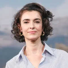 Blanca Romero, con su cabello rizado y pendientes dorados, sonríe con una camisa a rayas.