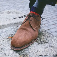 Zapato de gamuza marrón con cordones negros, calcetín a rayas y un consejo sobre como quitar manchas de humedad en zapatos de piel.