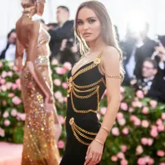 Lily-Rose Depp deslumbrante con un vestido negro adornado con cadenas doradas, posando ante un fondo de flores rosas y fotógrafos.