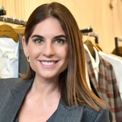 Lourdes Montes sonríe con un traje gris a cuadros, rodeada de ropa colgada en perchas en una tienda.