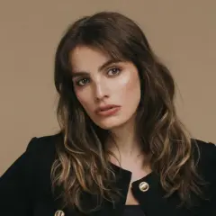 Retrato de una mujer con cabello castaño ondulado y flequillo, luciendo un blazer negro. Su mirada es intensa, transmitiendo un aire de misterio y elegancia, como si fuera la protagonista de una historia de lux pascal.