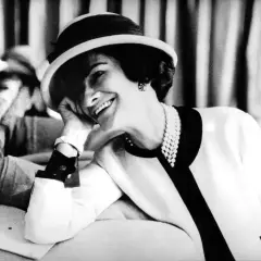 Coco Chanel, sonriendo con un sombrero y perlas, icónica figura de la moda.
