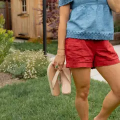 Mujer con blusa vaquera y shorts rojos, sosteniendo chanclas. El jardín tiene césped y flores.