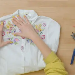 Manos embelleciendo una camisa blanca con bordados florales, lista para un proyecto de upcycling ropa. Herramientas de costura a un lado.