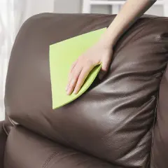 Mano limpiando un sofá de cuero marrón con una esponja verde para quitar manchas de desteñido de cuero.