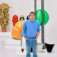 Jonathan Anderson posa entre esculturas y objetos de diseño, incluyendo una figura de madera tallada y un gran globo verde.