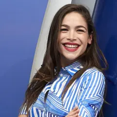Cosima Ruiz de la Prada, sonriendo con labios rojos y camisa a rayas azules y blancas, se apoya en una pared azul.