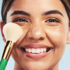 Una mujer sonríe mientras se aplica maquillaje con una brocha. Descubre trucos de maquillaje profesional para realzar tu belleza natural.
