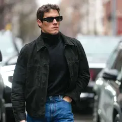 Hombre con estilo casual, viste jersey negro de cuello alto, chaqueta de ante y jeans. Camina por la calle entre coches.