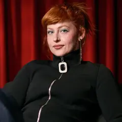 Chloe Wallace, con su cabello pelirrojo recogido y maquillaje azul, posa frente a una cortina roja.