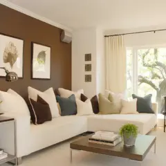 Sofá blanco con cojines en tonos tierra, azul y marrón. La pared marrón crea un ambiente acogedor, mostrando cómo los colores que combinan con marrón pueden ser elegantes.