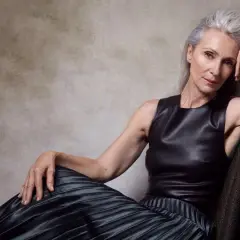 Mujer mayor con cabello plateado, vestida con un top de cuero y falda plisada, posa con elegancia. Su mirada es serena, como un pino montesdeoca en la cima de una montaña.