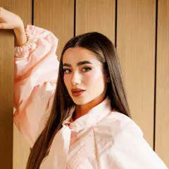 Lola Lolita, con su cabello largo y liso, posa con un vestido rosa pálido, apoyando su mano en un pilar de madera.