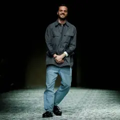 Matthieu Blazy, diseñador de Bottega Veneta, sonríe en la pasarela con camisa gris y vaqueros.