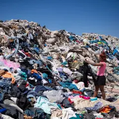 Montaña de ropa desechada, reflejo de las **fast fashion consecuencias**. Una joven busca entre los residuos textiles bajo un cielo azul.