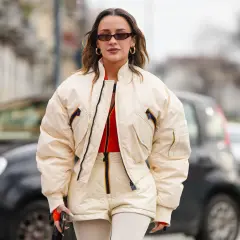 Mujer con gafas de sol y pendientes dorados, luciendo una bomber acolchada color crema sobre un top naranja y shorts a juego.