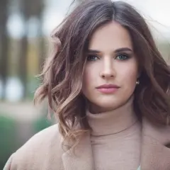 Mujer con una elegante media melena ondulada, ojos verdes y labios rosados, vestida con un abrigo beige.