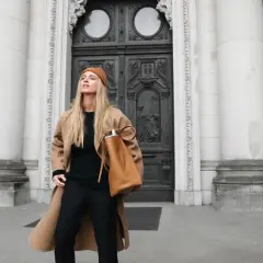 Mujer con abrigo camel, gorro y bolso marrón, luciendo un elegante pantalon negro.