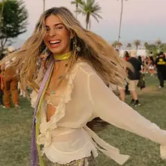 Mujer sonriente y con el pelo al viento disfruta de un look festival verano, rodeada de palmeras y gente.