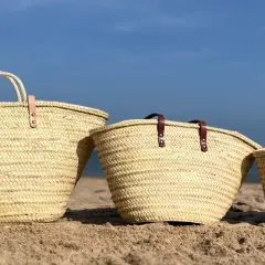 Tres capazos de playa grandes de palma, con asas de cuero, descansan en la arena bajo un cielo azul.