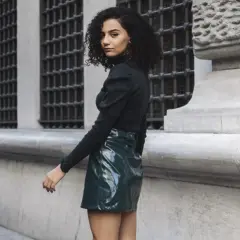 Joven con pelo rizado y falda verde brillante, estilo inspirado en Mary Quant, posa frente a un edificio histórico.