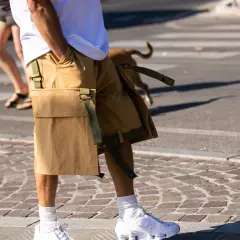 Hombre con shorts beige, camiseta blanca y zapatillas deportivas blancas, paseando por la calle con un perro.