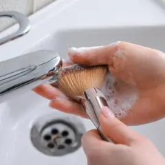 Manos limpiando brochas de maquillaje con espuma y agua bajo un grifo cromado en un lavabo blanco.