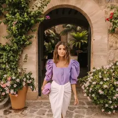 Mujer con ropa que combina color lila claro, un top abullonado y falda blanca, paseando entre vegetación exuberante.