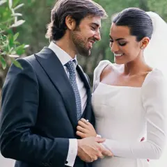 Marta Lozano y su esposo, radiantes en su boda, comparten una mirada cómplice y sonrisas. Él, con traje oscuro y corbata, la abraza tiernamente.