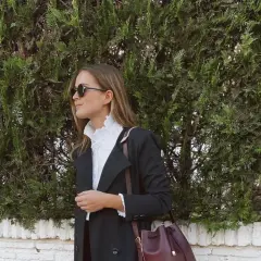 Mujer con elegante outfit para Semana Santa, abrigo negro, blusa blanca con volantes y bolso burdeos.