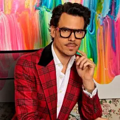 Juan Avellaneda, con su característico traje a cuadros rojos y gafas llamativas, posa con una mano en la barbilla frente a un vibrante cuadro abstracto.