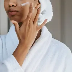 Mujer con bata blanca y toalla en la cabeza aplicándose crema en la mejilla, un gesto de cuidado para la piel con colágeno.