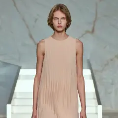 Modelo con vestido plisado color melocotón, en un escenario minimalista con escaleras blancas y fondo de mármol.