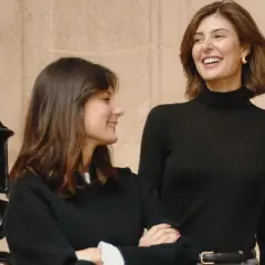 María de la Orden y su amiga ríen juntas frente a una reja ornamental.