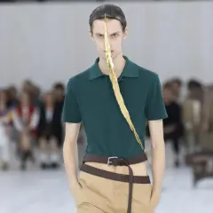 Enzo Vogrincic, modelo con un polo verde, pantalones caqui y un adorno dorado en la cara, desfila ante un público difuminado.