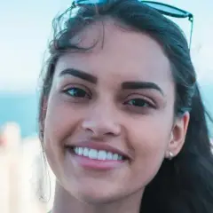 Sonrisa radiante de una joven con rostro ovalado, cabello oscuro y gafas de sol en la cabeza. Fondo de ciudad y mar.