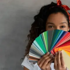 Mujer con cabello rizado y diadema roja sostiene un abanico de muestras de pintura, creando un efecto de color block.
