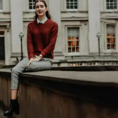 Chica con estilos de ropa modernos: jersey de punto rojo, camisa blanca, pantalones de cuadros y botines negros, sentada frente a un edificio clásico.